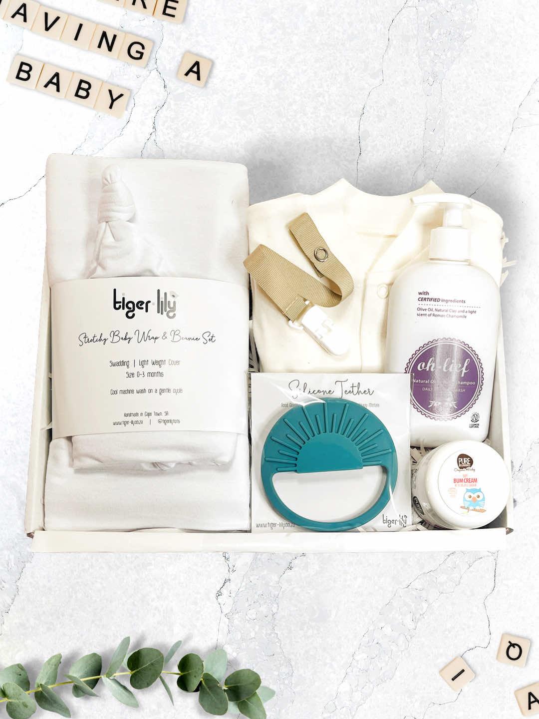 A baby shower gift set displayed on a white surface, including a baby grower, teether, dummy clip, shampoo, cream, and swaddling wraps.