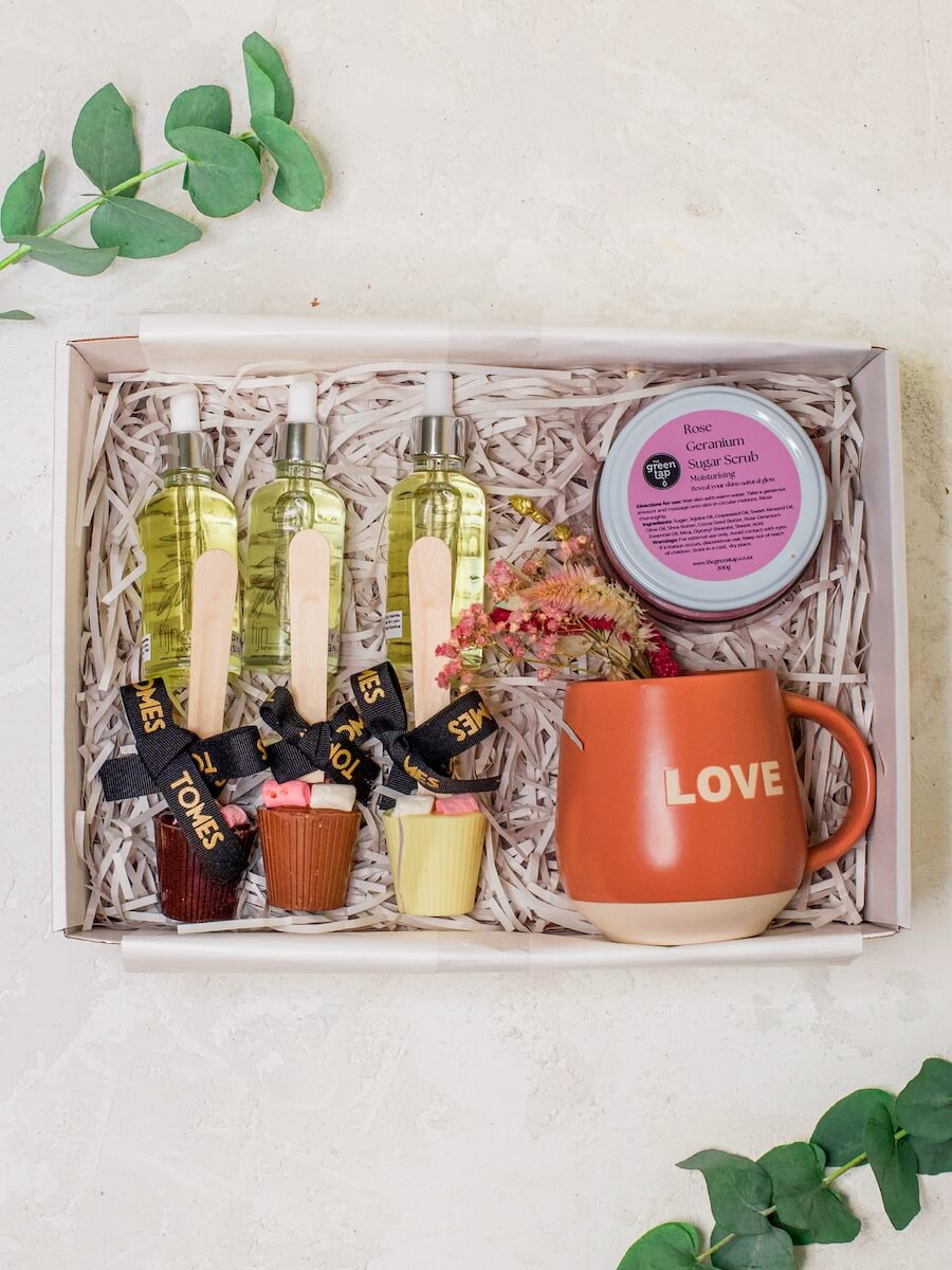 A gift set containing various skincare products and a mug with 'LOVE' written on it, presented in a wooden box lined with shredded paper.