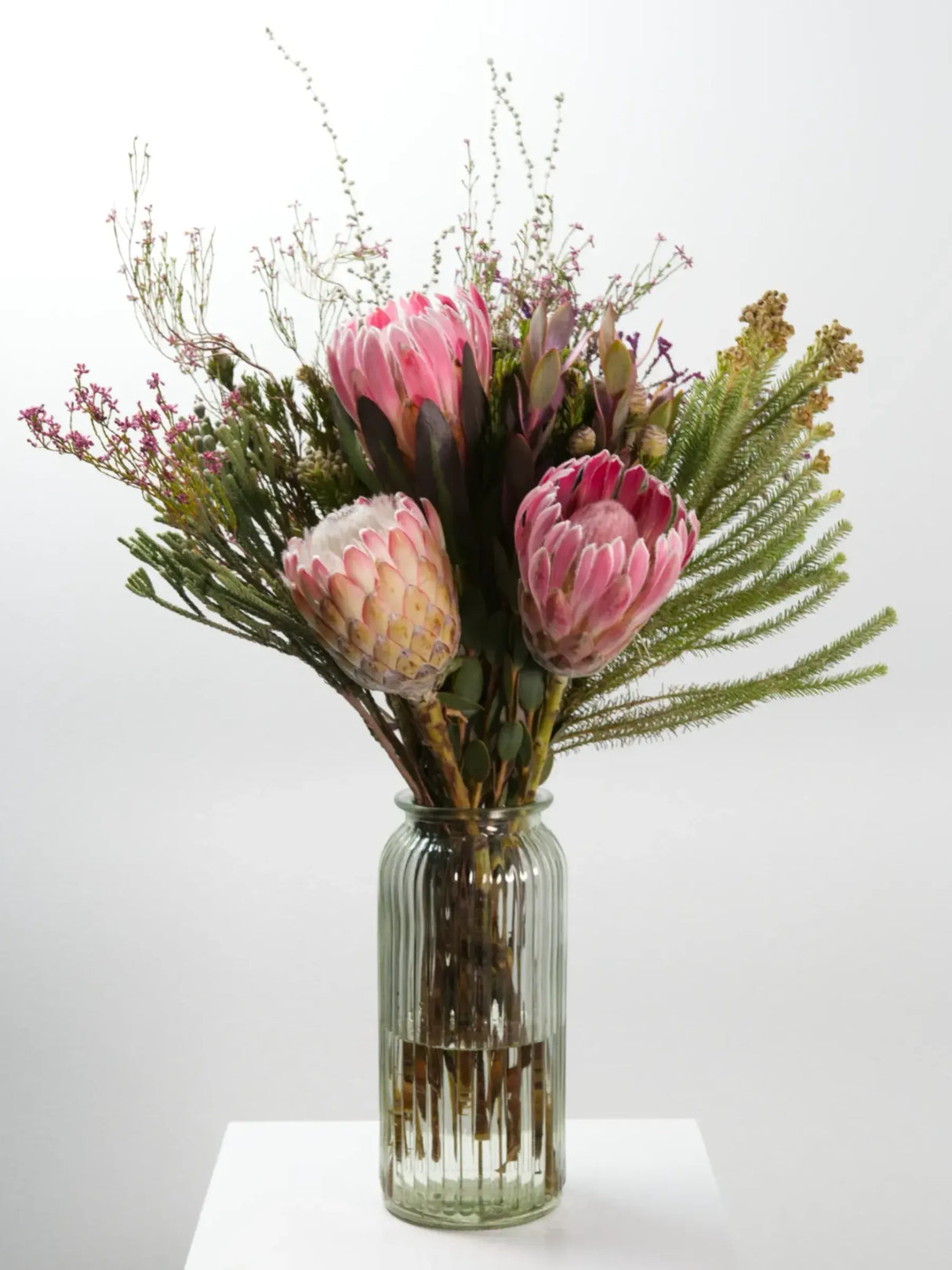 Bloomable's Girl You Is Fynbos arrangement in a textured glass bottle vase on white surface. Features beautiful pink king proteas, cream-pink blushing bride protea, feathery fern fronds, and delicate wispy filler flowers. This elegant South African native floral arrangement makes a striking gift for birthdays, housewarmings or special celebrations with same-day delivery across South Africa.