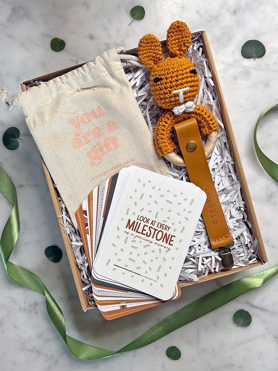 A gift box set on a white marble surface, featuring a cream drawstring pouch with pink text, a crocheted orange bunny keychain with a brown leather loop, a stack of white cards, and shredded paper inside a kraft box, with green ribbon and eucalyptus leaves around it.