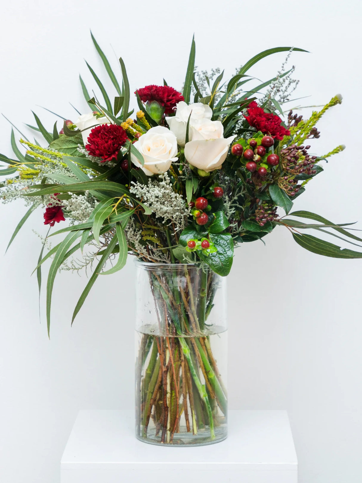 Natural Christmas flower arrangement featuring red carnations, cream roses, gypsophila, hypericum berries and mixed foliage in clear glass vase - rustic festive bouquet