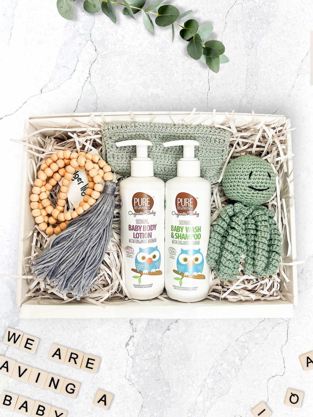 A gift box containing a hand crocheted baby toy, a beaded tassel, a crochet cotton washcloth, and bottles of organic baby wash and lotion.