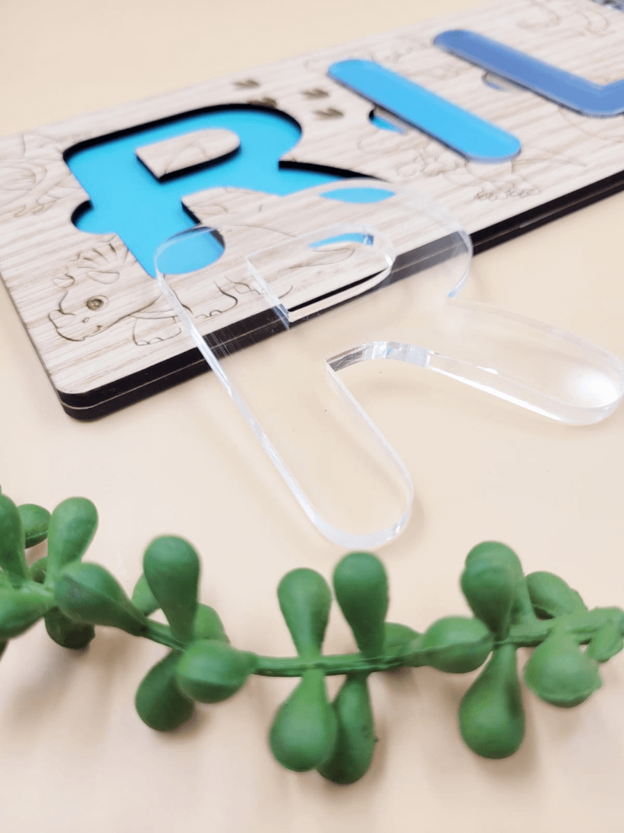 Personalised engraved name puzzle for kids with blue and clear acrylic letters on oak veneer, next to a green plant accent.