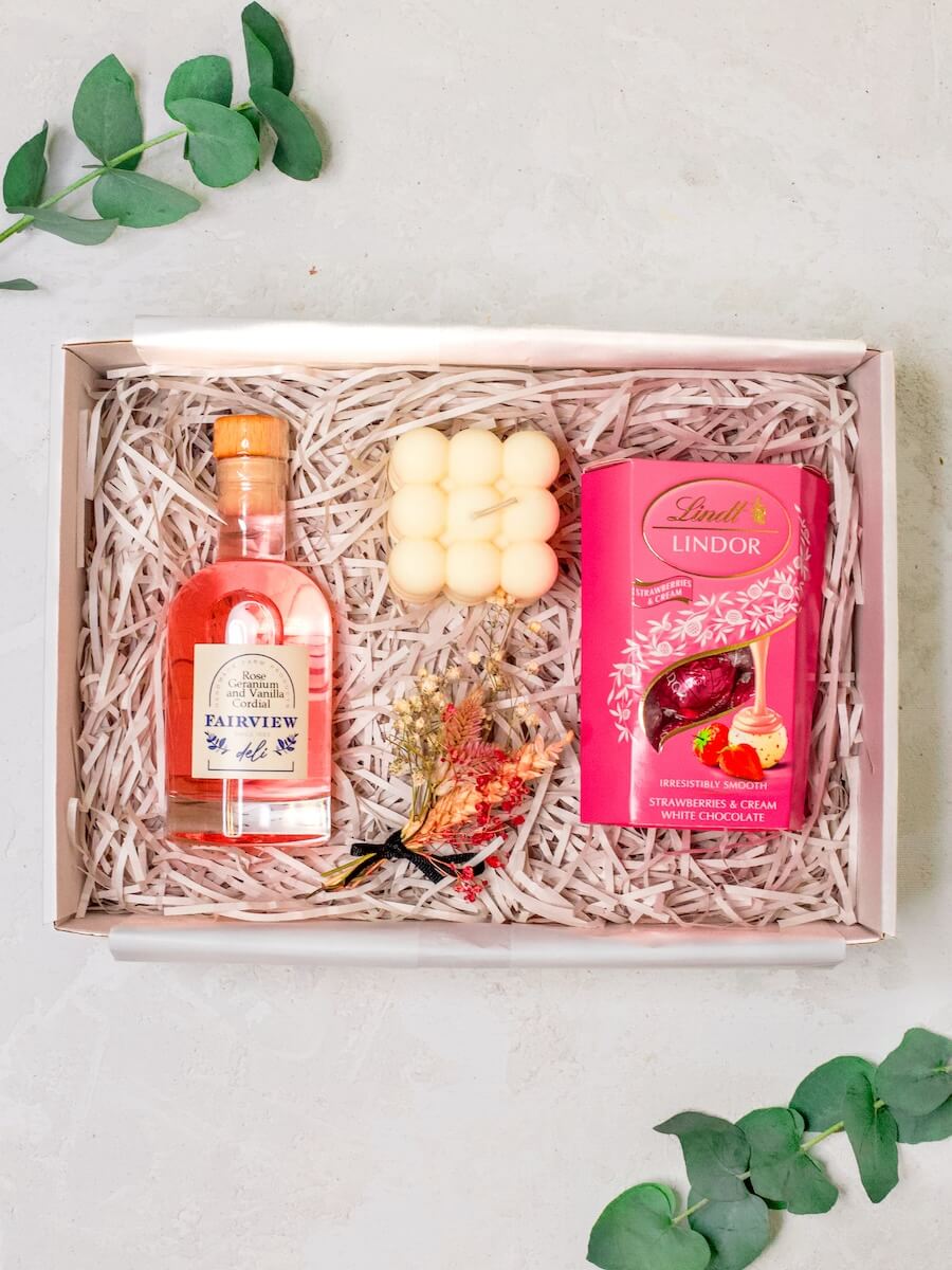 A gift box containing a pink soy wax candle, a bottle of Fairview Rose Geranium and Vanilla Cordial, a box of Lindt Lindor Strawberries & Cream Truffles, and a dried flower mini keepsake posy.