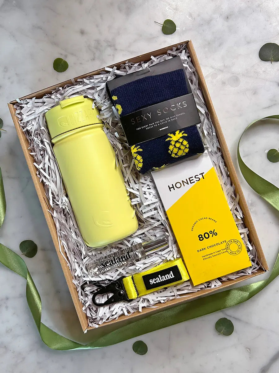 A curated gift box set on a marble surface, featuring a bright yellow insulated bottle, navy socks with yellow pineapples, a yellow Honest brand soap bar, and a yellow and black Sealand keychain, all arranged in white paper shred inside a wooden box.