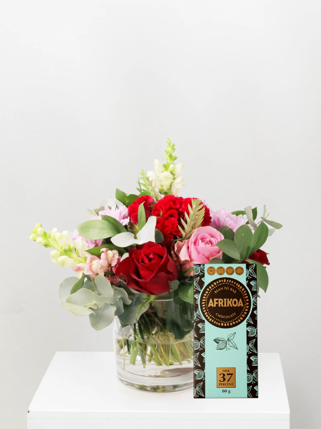 Bouquet of flowers in a glass vase next to a box of Afrikoa chocolate on a white surface.