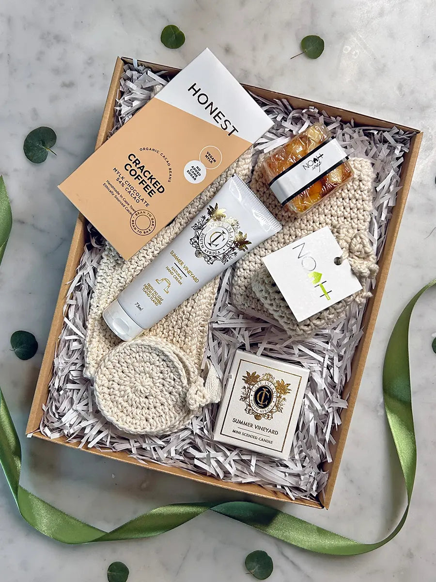 A curated gift box set on a light marble surface, containing a tube of Honest brand whipped body butter, a jar of honey, a cream-colored knit baby blanket, a round knit baby towel, a small tag, and a card from Domestically Sourced, all arranged in white shredded paper inside a brown box with a green ribbon.