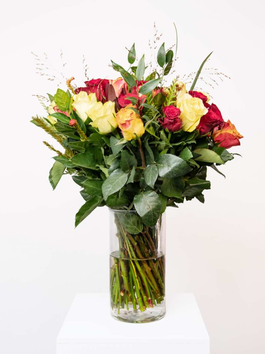Bouquet of mixed roses in red, orange, and yellow with lush greenery, arranged in a clear glass vase, perfect for expressing love and enthusiasm.