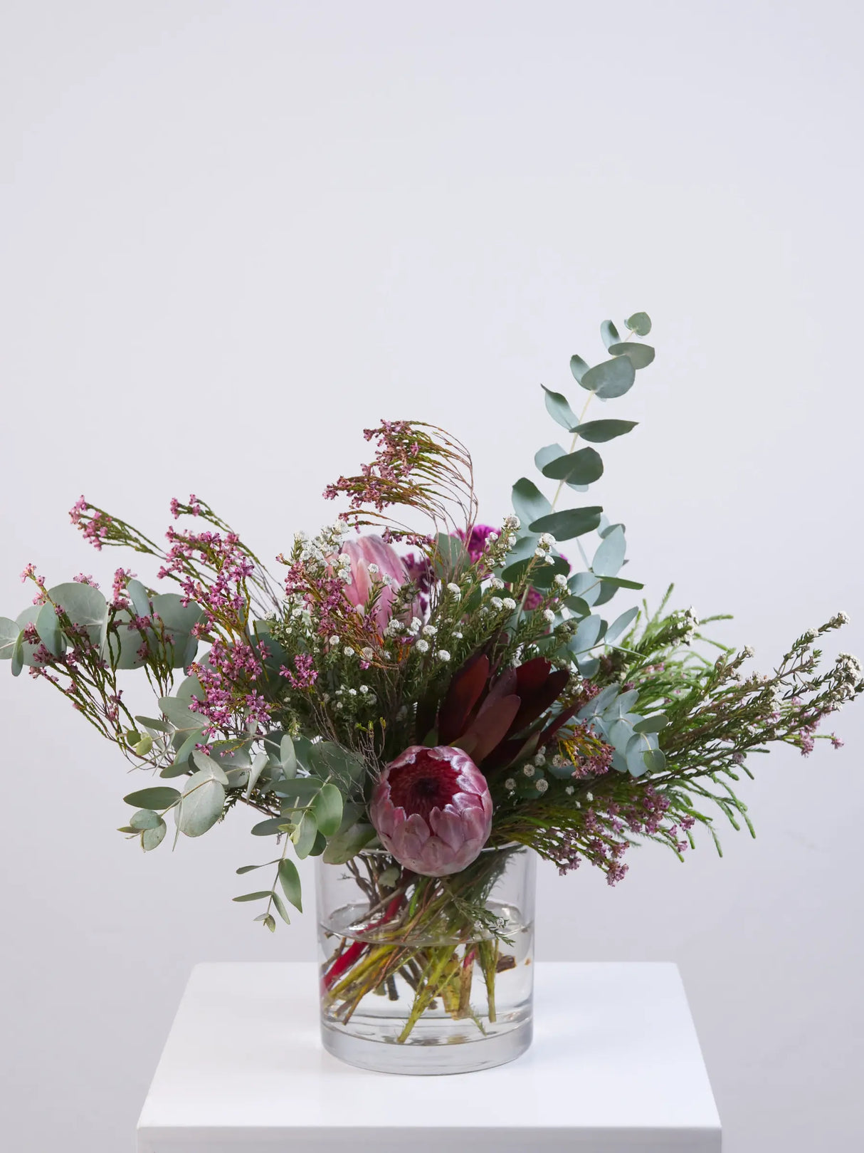 Bouquet of protoes and fynbos in a clear vase on a white surface with a light gray background