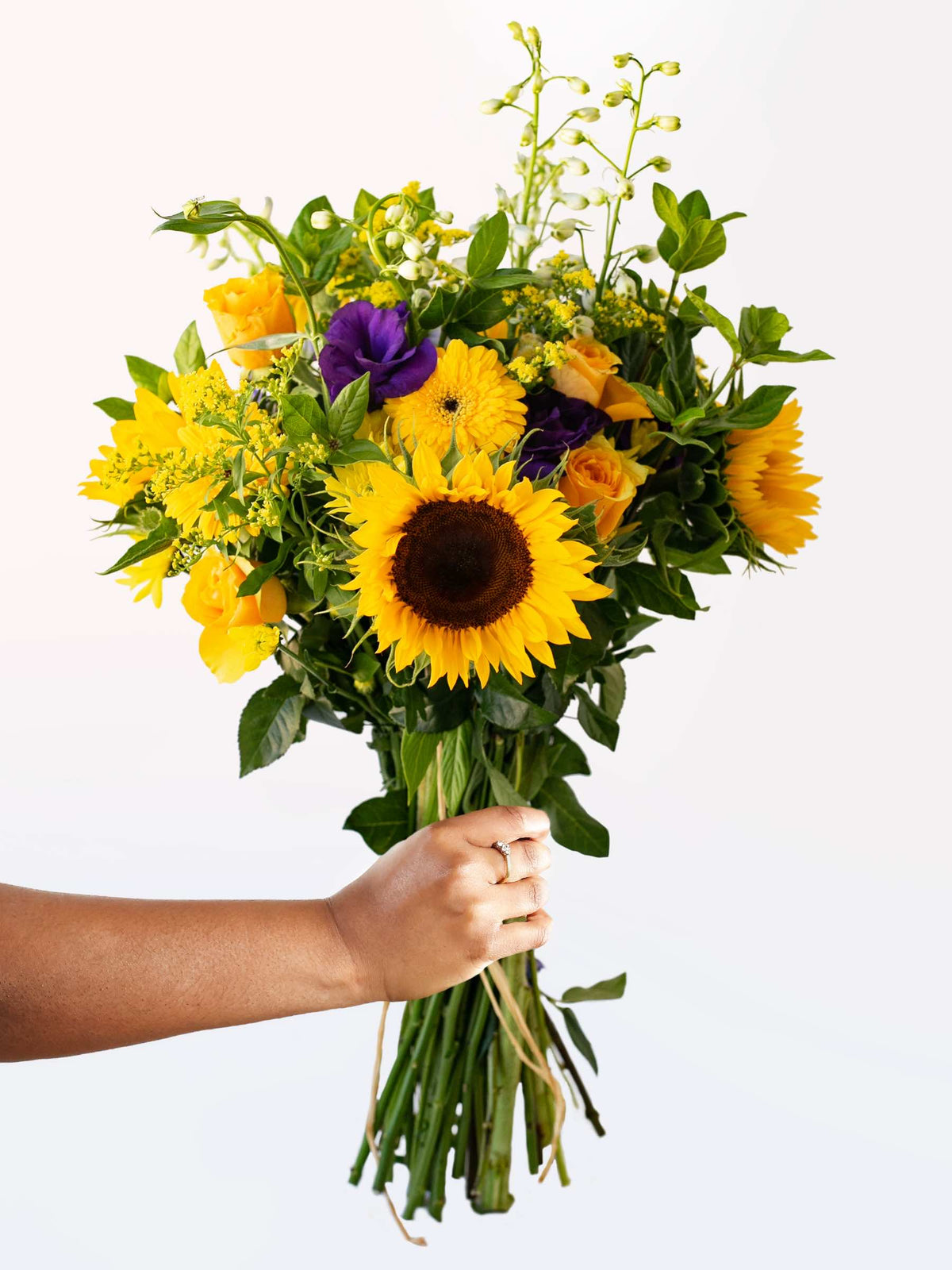 Hand holding a vibrant bouquet of seasonal purple and yellow flowers, featuring sunflowers and green foliage against a white background.