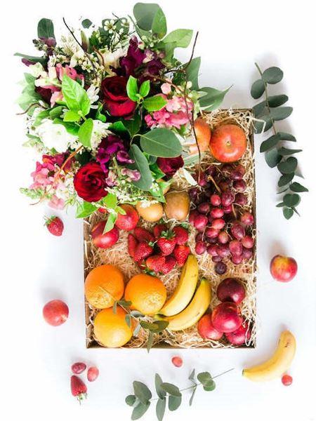 Gift arrangement of fresh fruits and vibrant flowers in a clear vase, including oranges, grapes, strawberries, nectarines, apples, and kiwi.