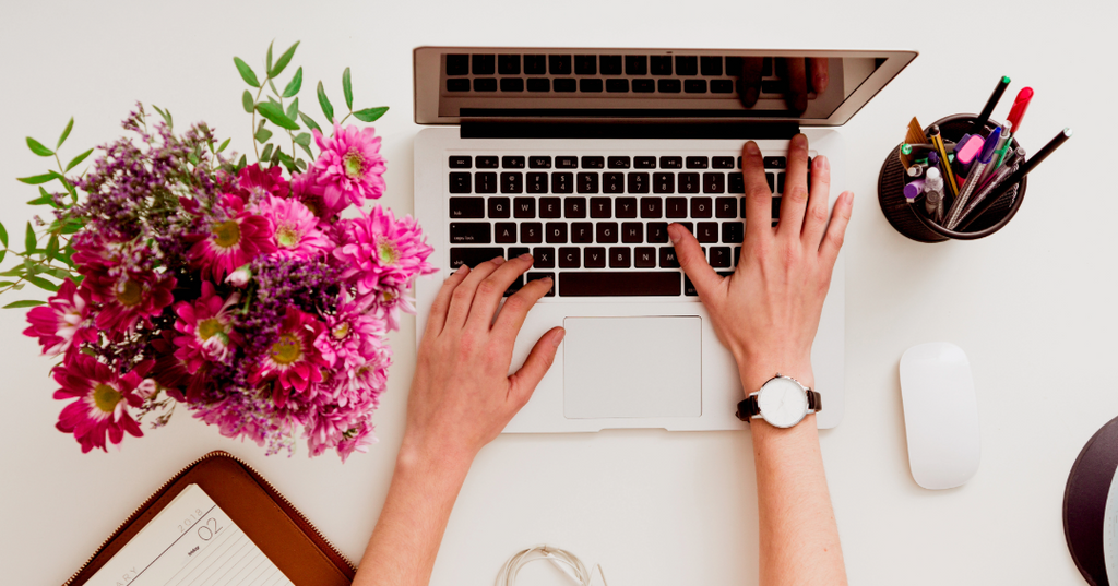 Desk with fresh flower arrangement and hands working on laptop with pens and notebook on desk 