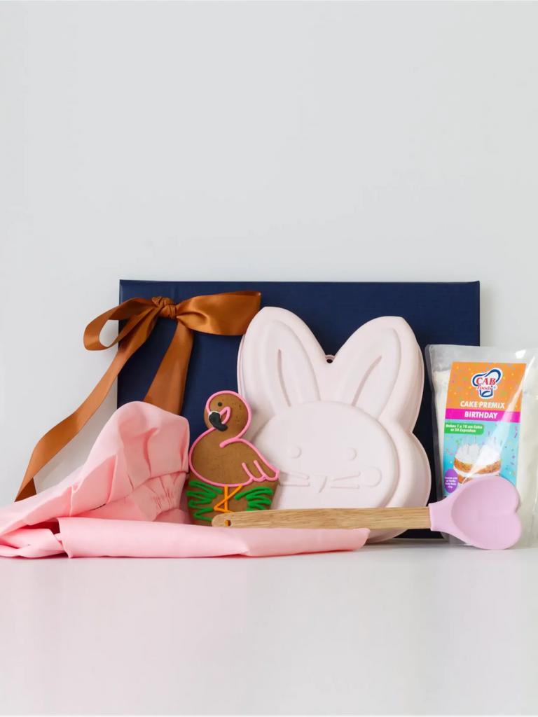 A pastel-themed baking gift set featuring a soft pink apron and baker’s hat, a pink silicone bunny-shaped baking mould, a vanilla cake mix, a pink heart-tipped spatula, and a beautifully decorated cookie from Hark & Heart, all styled in front of a navy blue gift box with copper ribbon.
