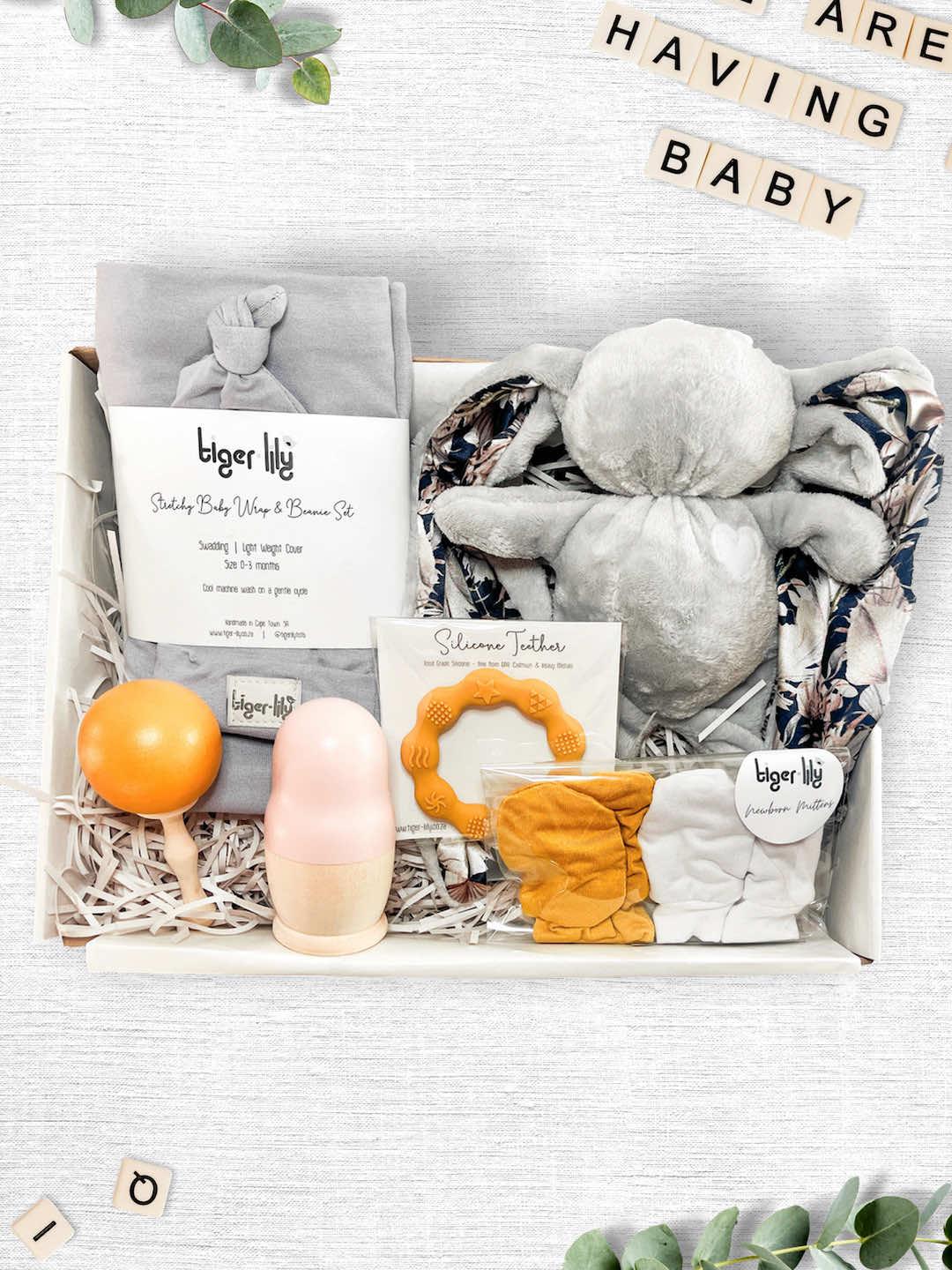 A luxury newborn baby gift box containing Tiger Lily brand items including a set of two white and gray striped newborn mittens, a wooden rattle, a set of nesting dolls, a silicone ring teether, and a gray baby wrap and beanie set, all displayed on a white background.