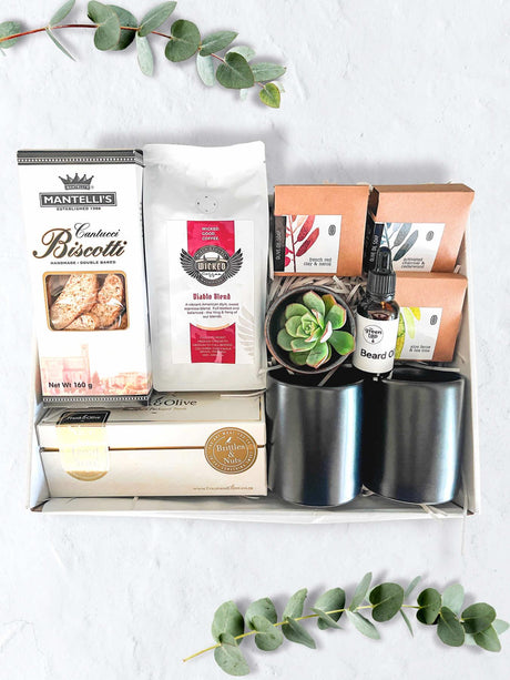 A luxury gift box for him containing beard oil, soap, handmade biscotti, coffee, a treat box with britles and nuts, and two black mugs, all displayed on a white surface with some greenery in the background.