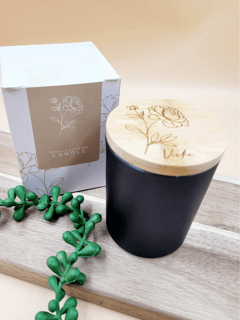 Personalised soy candle in black glass jar topped with engraved bamboo lid, alongside floral printed packaging. 