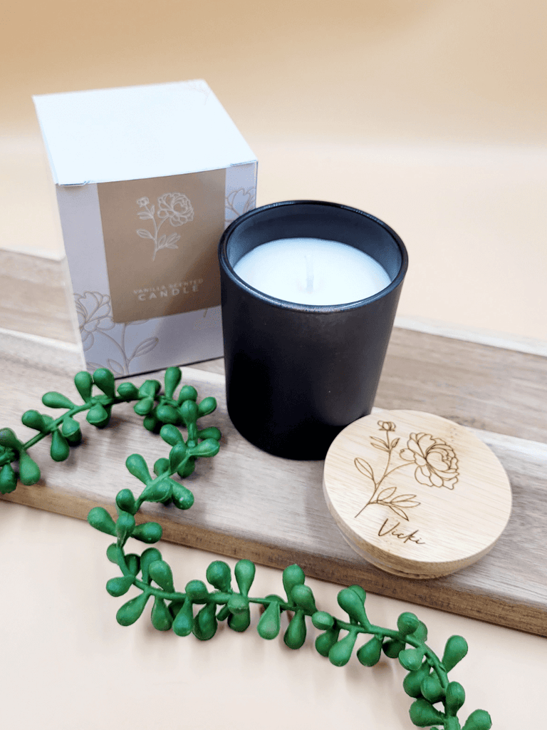 Personalised soy candle in black glass jar alongside engraved bamboo lid and floral printed packaging. 