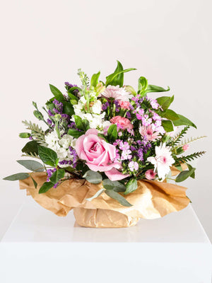 A beautiful floral arrangement featuring a variety of flowers in shades of pink, white, and green, presented in a round, low vase with brown paper wrap.