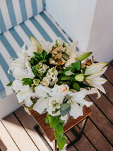Bouquet of white lilies and greenery on a wooden table with striped chair in the background