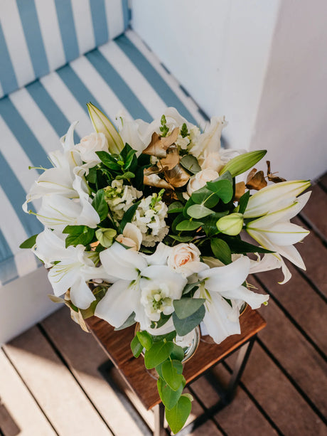 Bouquet of white lilies and greenery on a wooden table with striped chair in the background