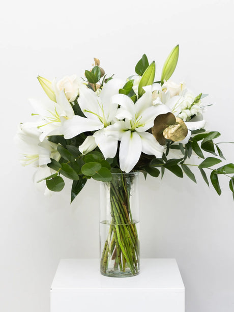 Bouquet of white lilies and greenery in a clear vase on a white surface with a white background