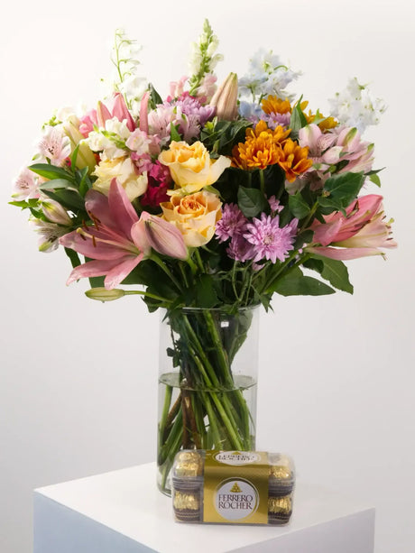 Bouquet of colorful flowers in a clear vase on a white surface with a white background with chocolates