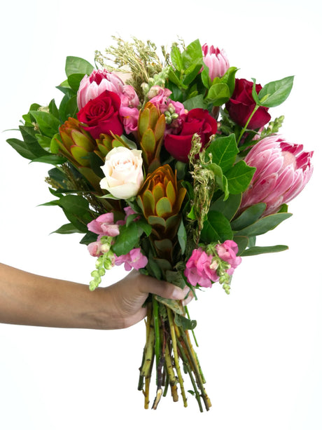 Hand holding bouquet of red roses, pink proteas, fynbos, and greenery in a stylish arrangement, highlighting romantic and whimsical vibes.