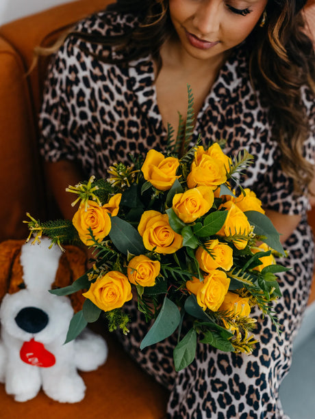Woman in leopard print dress holding a bouquet of yellow roses with a plush toy dog in the background.