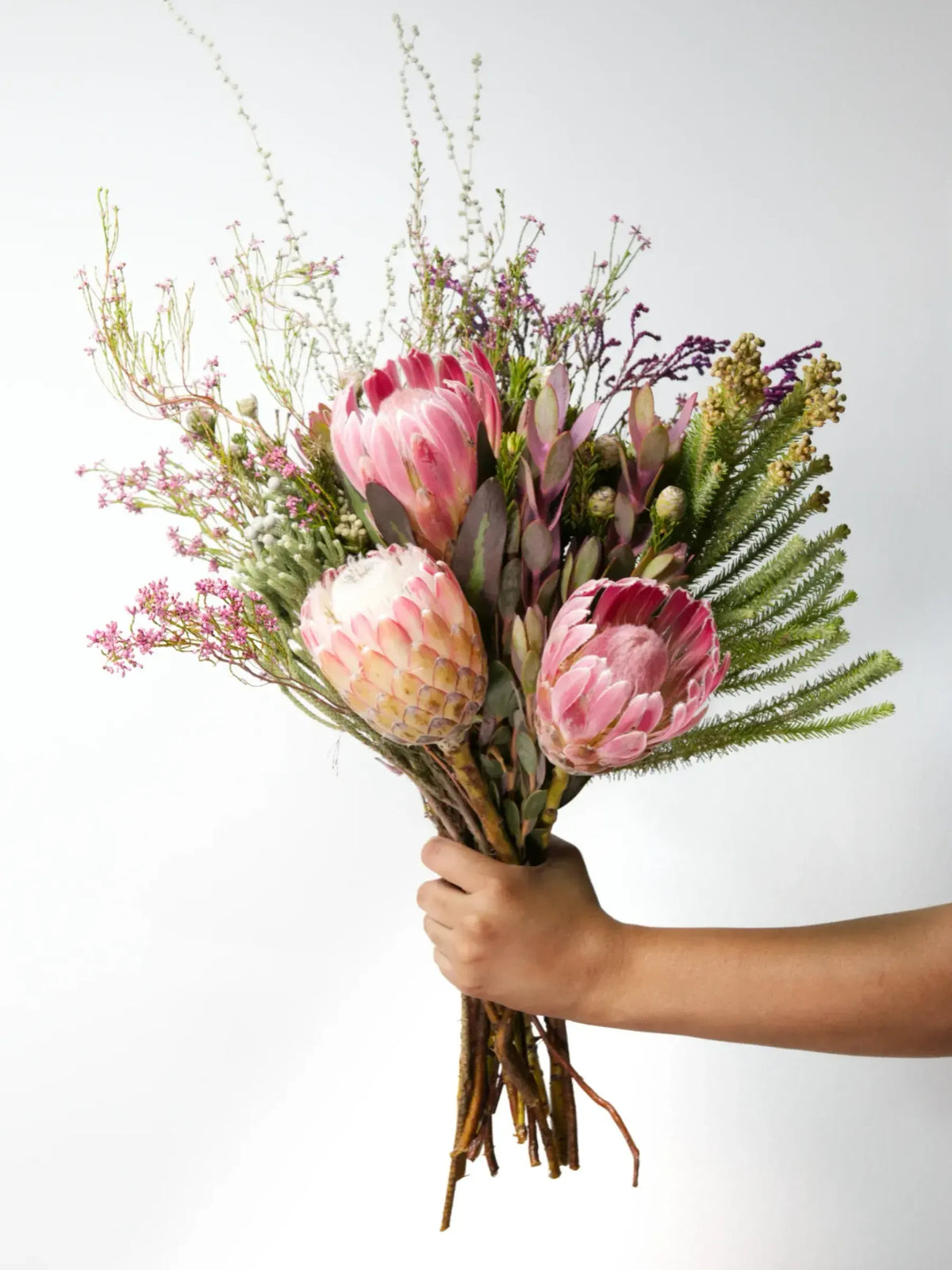 Bloomable's Girl You Is Fynbos hand-held bouquet featuring soft pink king proteas, cream-pink blushing bride protea, feathery fern fronds, and delicate pink filler flowers with wispy natural elements. This artfully arranged selection of South African native flora makes an elegant gift for Mother's Day, birthdays, anniversaries, or to show appreciation with same-day delivery across South Africa.