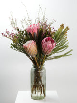 Bloomable's Girl You Is Fynbos arrangement in a textured glass bottle vase on white surface. Features beautiful pink king proteas, cream-pink blushing bride protea, feathery fern fronds, and delicate wispy filler flowers. This elegant South African native floral arrangement makes a striking gift for birthdays, housewarmings or special celebrations with same-day delivery across South Africa.