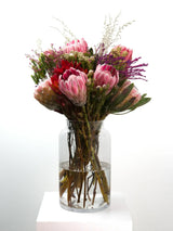 "Bloomable's Girl You Is Fynbos protea bouquet showcased in a modern cylindrical glass vase on a white plinth. Features an eye-catching collection of pink king proteas, vibrant red pincushions, delicate pink filler flowers, and textural greenery with natural woody stems. An exquisite South African fynbos arrangement ideal for Women's Day celebrations or stunning home décor with same-day delivery across South Africa."