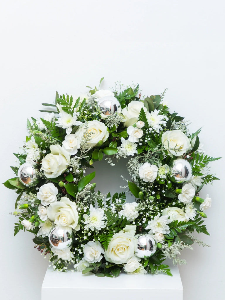 Elegant white and green floral wreath with roses, baby’s breath, and silver baubles — luxury flower arrangement for Christmas or memorial décor.