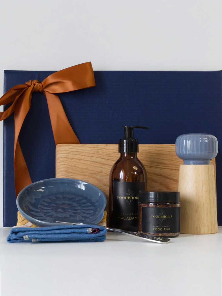 Displayed in front of the gift box is an oak serving board, and a hand-glazed blue ceramic side plate resting on a patterned cotton tea towel. Gourmet food items include Extra Virgin Macadamia Oil and a Fennel & Citrus Food Rub, both branded "FOODPHORIA." Completing the set is a practical and elegant salt grinder with a wooden base. 