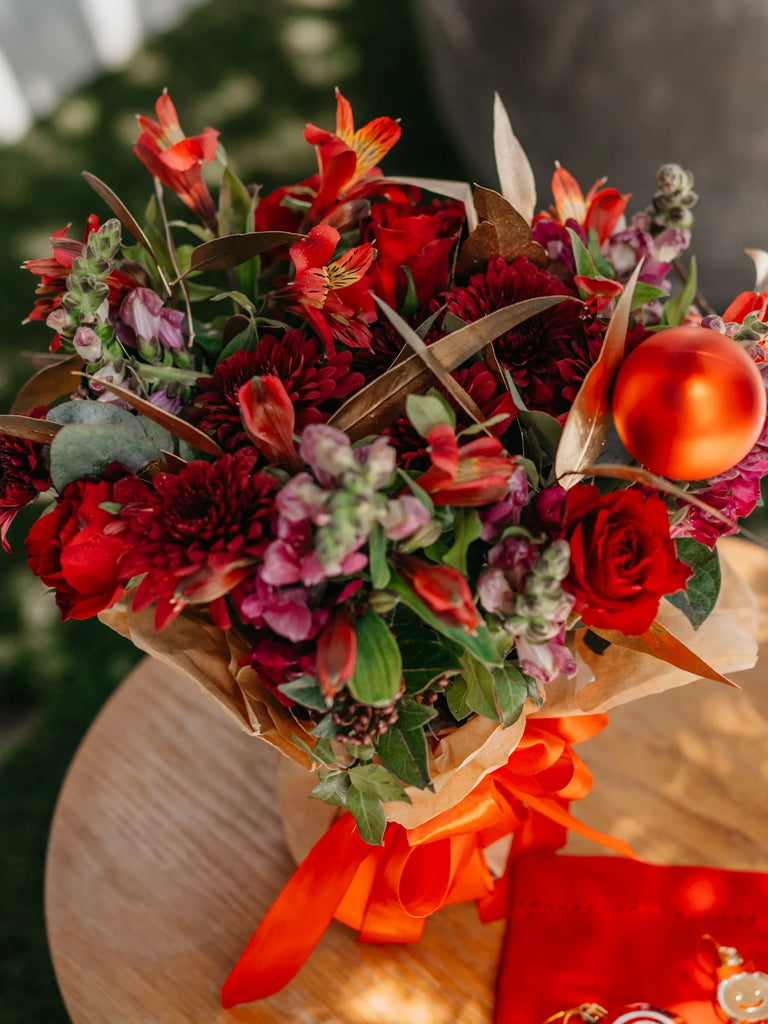Christmas flower bouquet gift with red roses, burgundy blooms, purple stocks and red bauble held outdoors - festive gift delivery