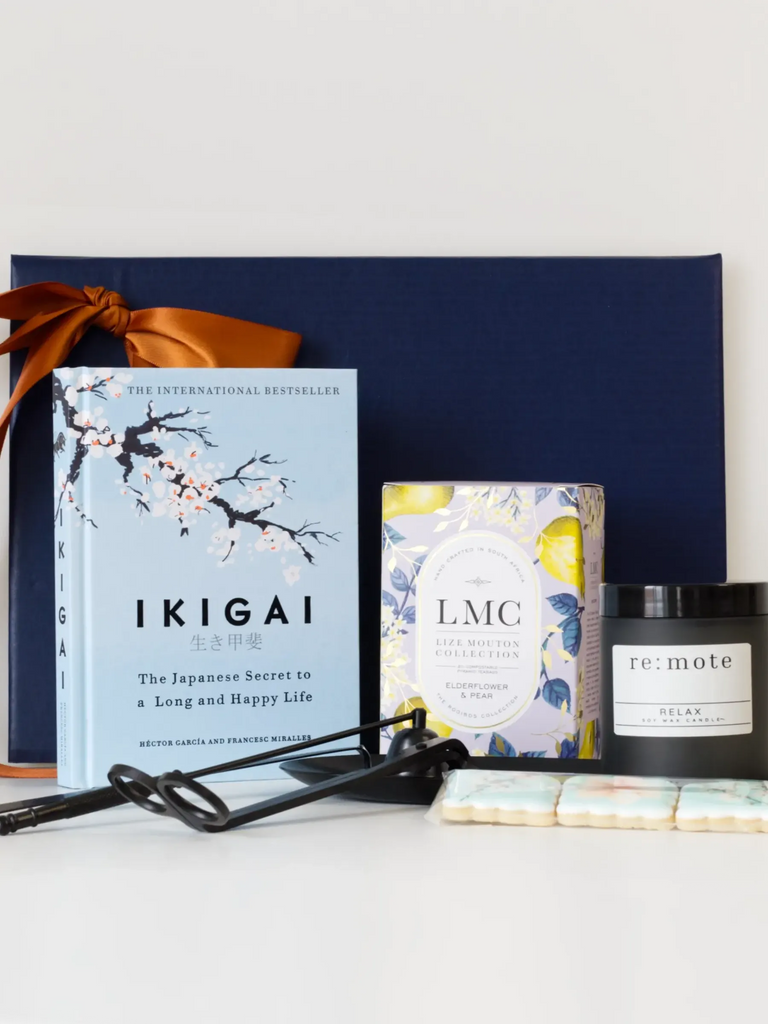 A beautifully arranged gift set featuring the book Ikigai: The Japanese Secret to a Long and Happy Life, an LMC Elderflower & Pear tea box with floral packaging, a black re: mote “Relax” soy candle, a set of matte black candle tools with a matching tray, and a row of pastel-decorated shortbread biscuits, all styled against a navy blue gift box with a copper satin ribbon.