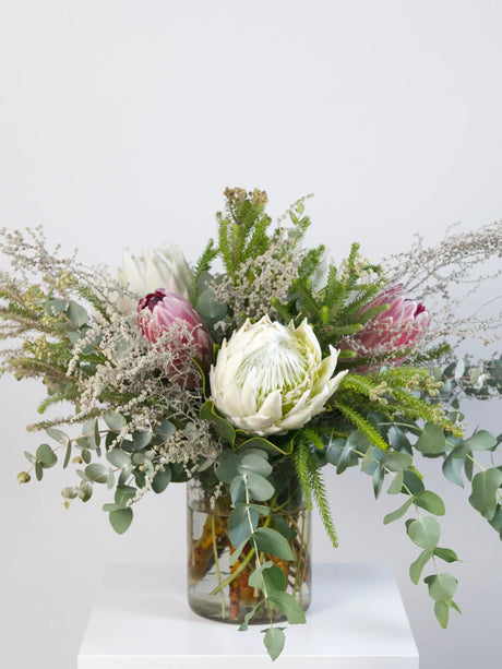 Floral arrangement with protea and eucalyptus in a clear vase on a white surface.