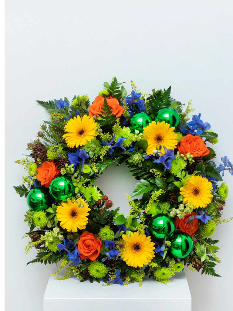 Colourful Christmas wreath with yellow gerbera daisies, orange roses, blue delphiniums, green chrysanthemums, and metallic baubles - vibrant festive door decoration