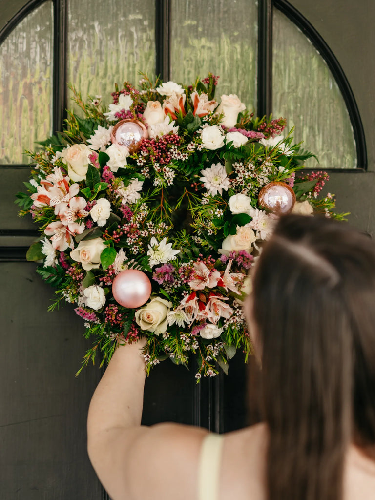 Luxury blush pink Christmas wreath with cream roses, baby's breath, evergreen foliage, rose gold baubles and berries - festive door decoration held by woman
