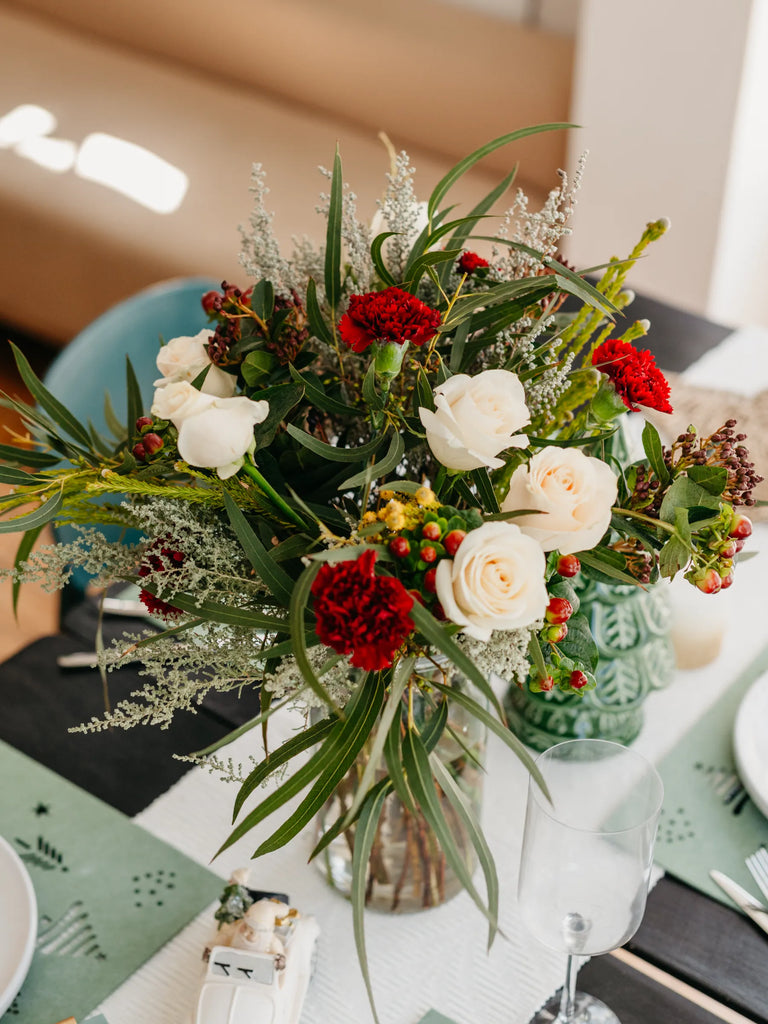 Christmas flower bouquet featuring cream roses, red carnations, baby's breath and hypericum berries in clear vase - festive desk arrangement for home office or workplace