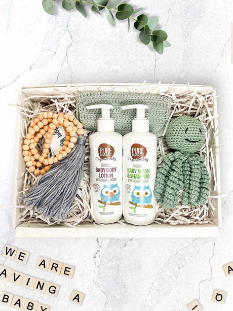 A gift box containing a hand crocheted baby toy, a beaded tassel, a crochet cotton washcloth, and bottles of organic baby wash and lotion.