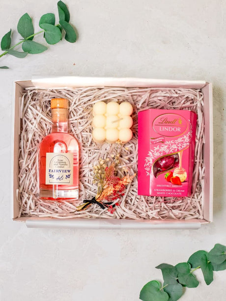 A gift box containing a pink soy wax candle, a bottle of Fairview Rose Geranium and Vanilla Cordial, a box of Lindt Lindor Strawberries & Cream Truffles, and a dried flower mini keepsake posy.