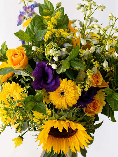 Colorful bunch of seasonal flowers in blues and yellows with sunflowers, roses, and foliage in a vibrant arrangement.