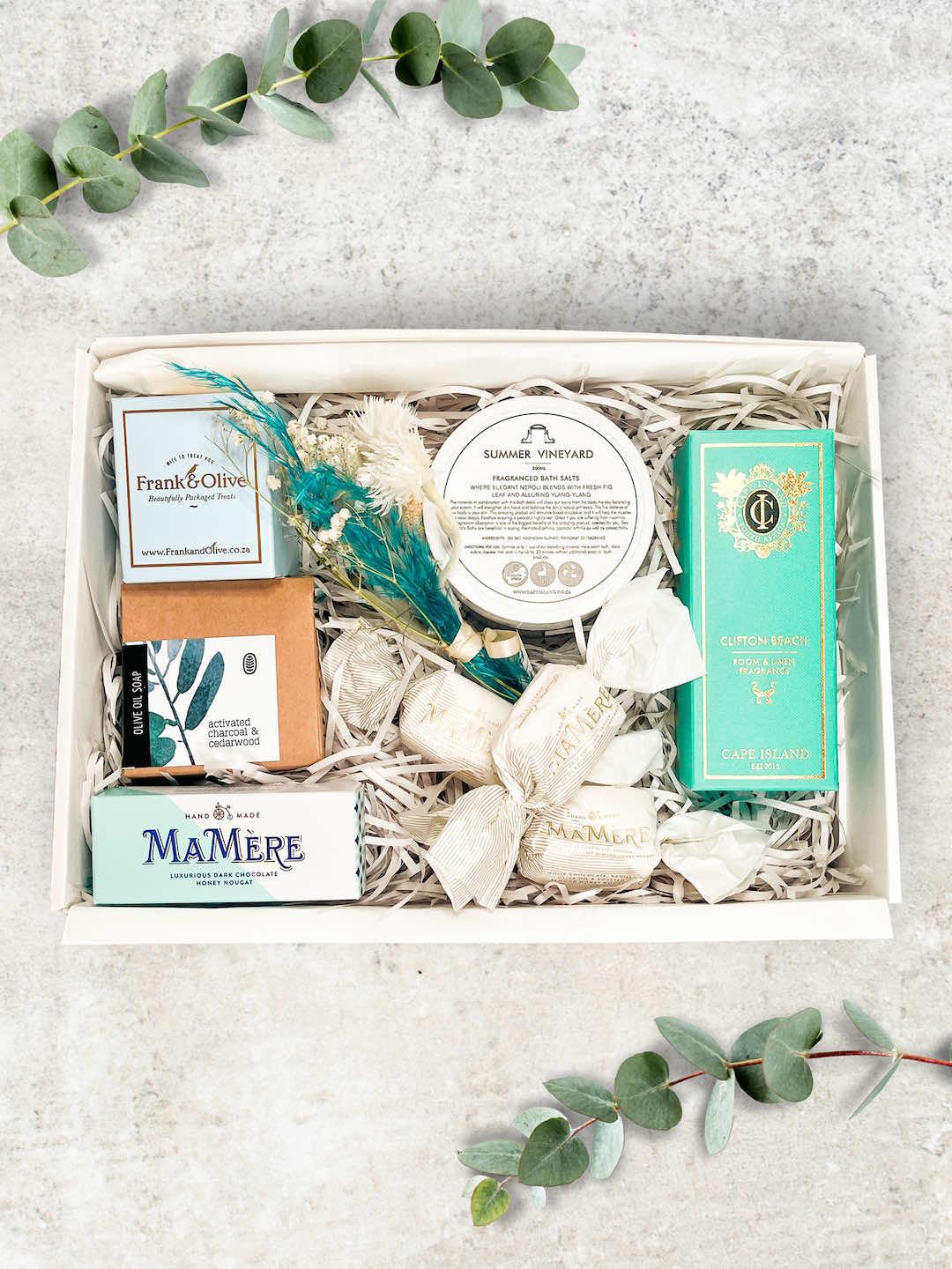 A gift basket containing bath salts, a room spray, olive oil soap, and chocolate nougat snacks, all arranged neatly in a box.