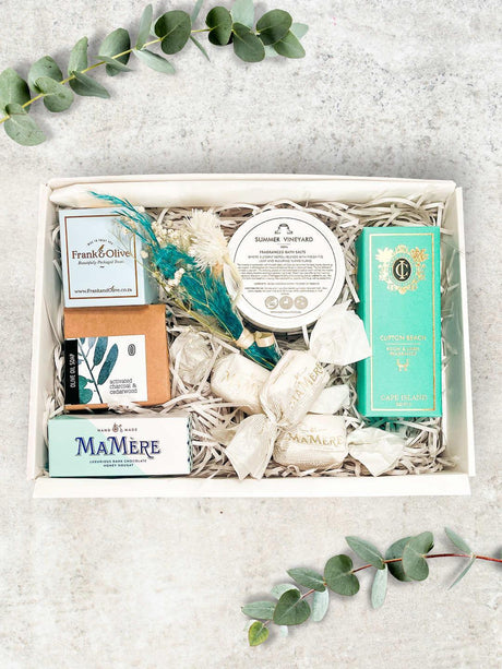 A gift basket containing bath salts, a room spray, olive oil soap, and chocolate nougat snacks, all arranged neatly in a box.