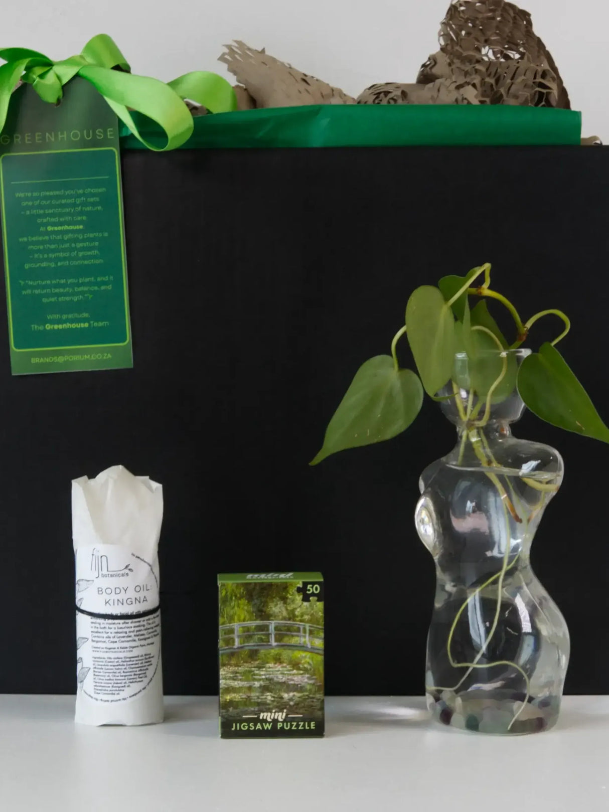 A gift set featuring a black box with a green ribbon and tag, a bottle of Fijn Kingra body oil, a mini jigsaw puzzle box depicting Monet's Water Lily Pond, and a small Pothos plant with roots visible in a clear glass female-torso-shaped vase.