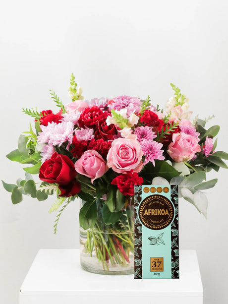 Bouquet of red and pink flowers in a clear vase with a bar of chocolate labeled 'AFRIKOA' on a white surface.