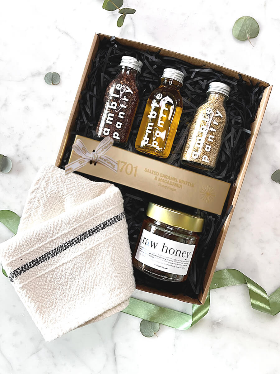 Gift box of gourmet delights with olive oil, dukkah, rooibos, honey, caramel brittle, nougat, and a tea towel, perfect for gifting.