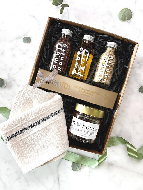 Gift box of gourmet delights with olive oil, dukkah, rooibos, honey, caramel brittle, nougat, and a tea towel, perfect for gifting.