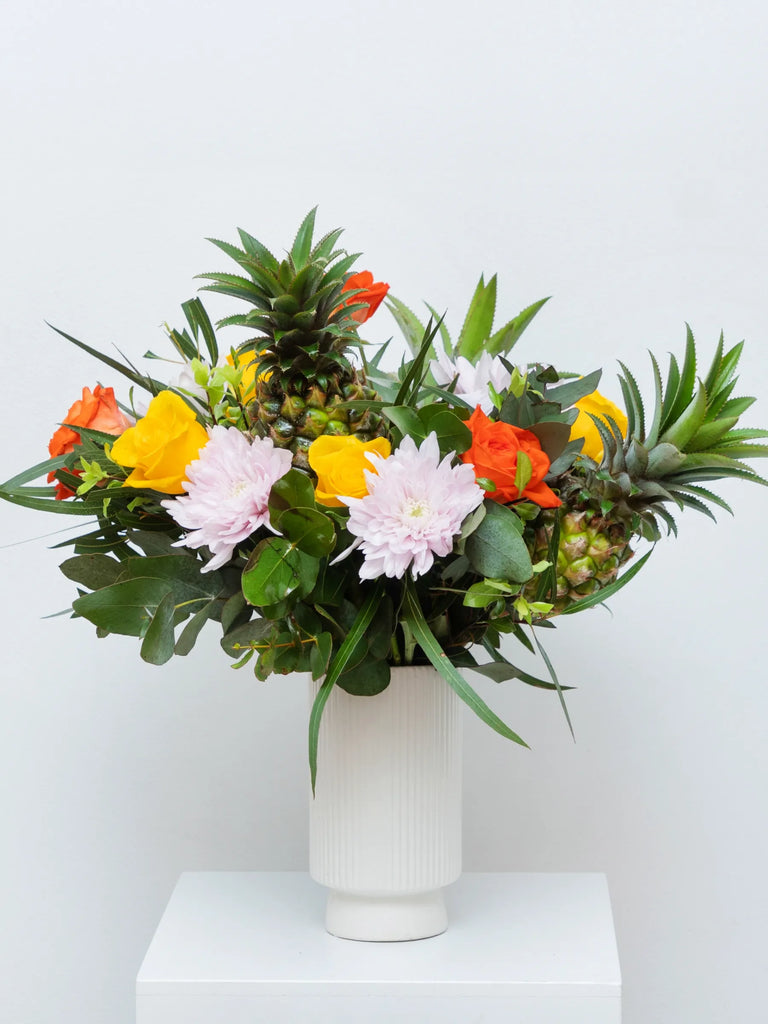 Tropical flower arrangement with fresh pineapples, white chrysanthemums, orange and yellow flowers in white ceramic vase - exotic centrepiece
