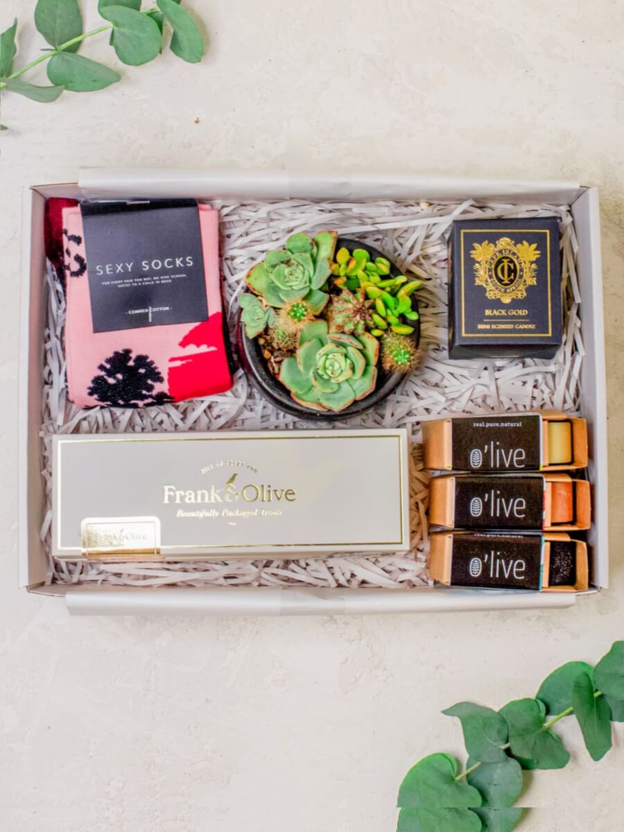 A gift set for him containing Locally Crafted O'Live Artisanal Soaps, a potted succulent mini garden, Frank & Olive Brittles & Nuts Box, a pair of Sexy Socks African Sunset, and a Black Gold Mini Scented Candle by Cape Island, all arranged in a luxurious gift box.